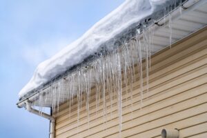 Winter Roof Damage. Sharp Spiked Icicles On Roof With Snow Against Blue Sky.