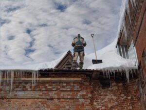 Commercial Roofing. Man, cleans the roof of a commercial building from snow with a shovel, The concept of the threat of falling ice and icicles. Municipal service.