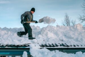 Winter Roof Repairs. Caucasian male with shovel remove snow from roof of totally snow covered house.